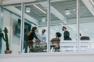 A modern office meeting room with glass walls. Inside, four professionals appear engaged in a discussion. Two men and two women: one man is standing and gesturing, actively speaking to the group. A woman, also standing, appears to be in conversation with him. The other man and woman are sitting and seem to be listening attentively. The room features a simple interior with a cactus plant, a wall-mounted monitor, and a few chairs around a small meeting table. Natural light filters in from the ceiling, creating a bright and airy atmosphere.